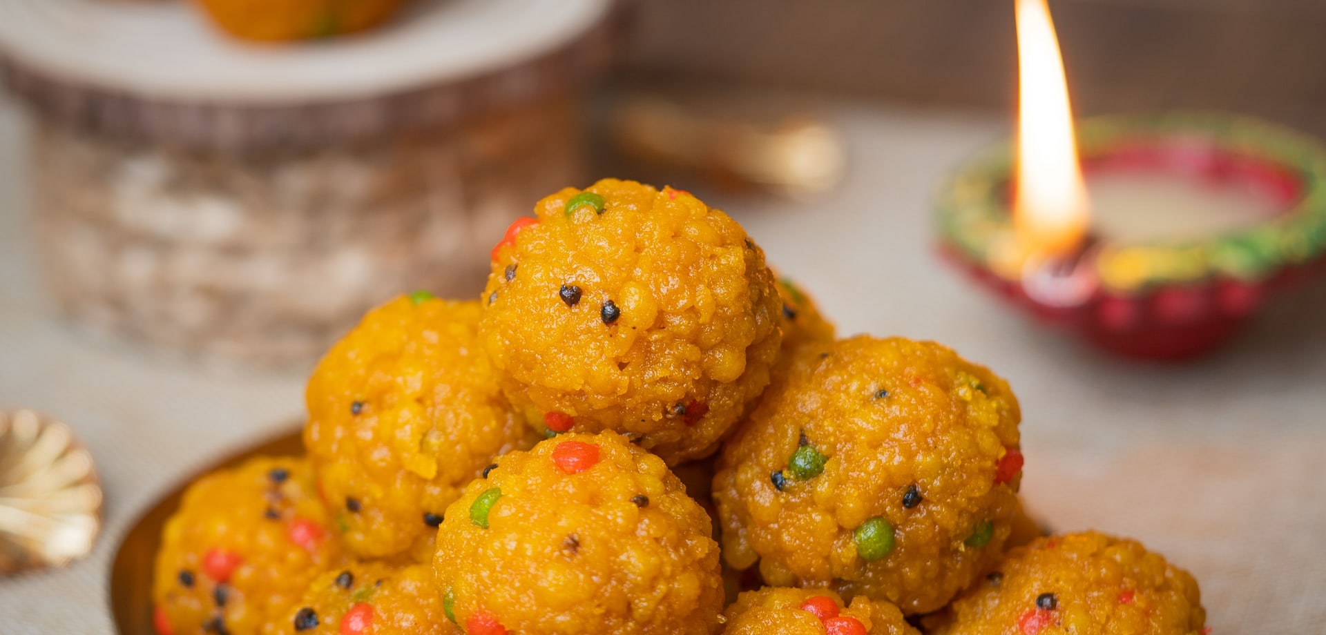 A plate of boondi laddoos beside a lit clay diya, on a home kitchen table